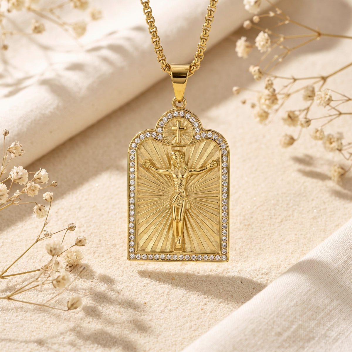 Close-up of a gold-tone arched crucifix pendant with sunburst rays and pavé accents above sand.