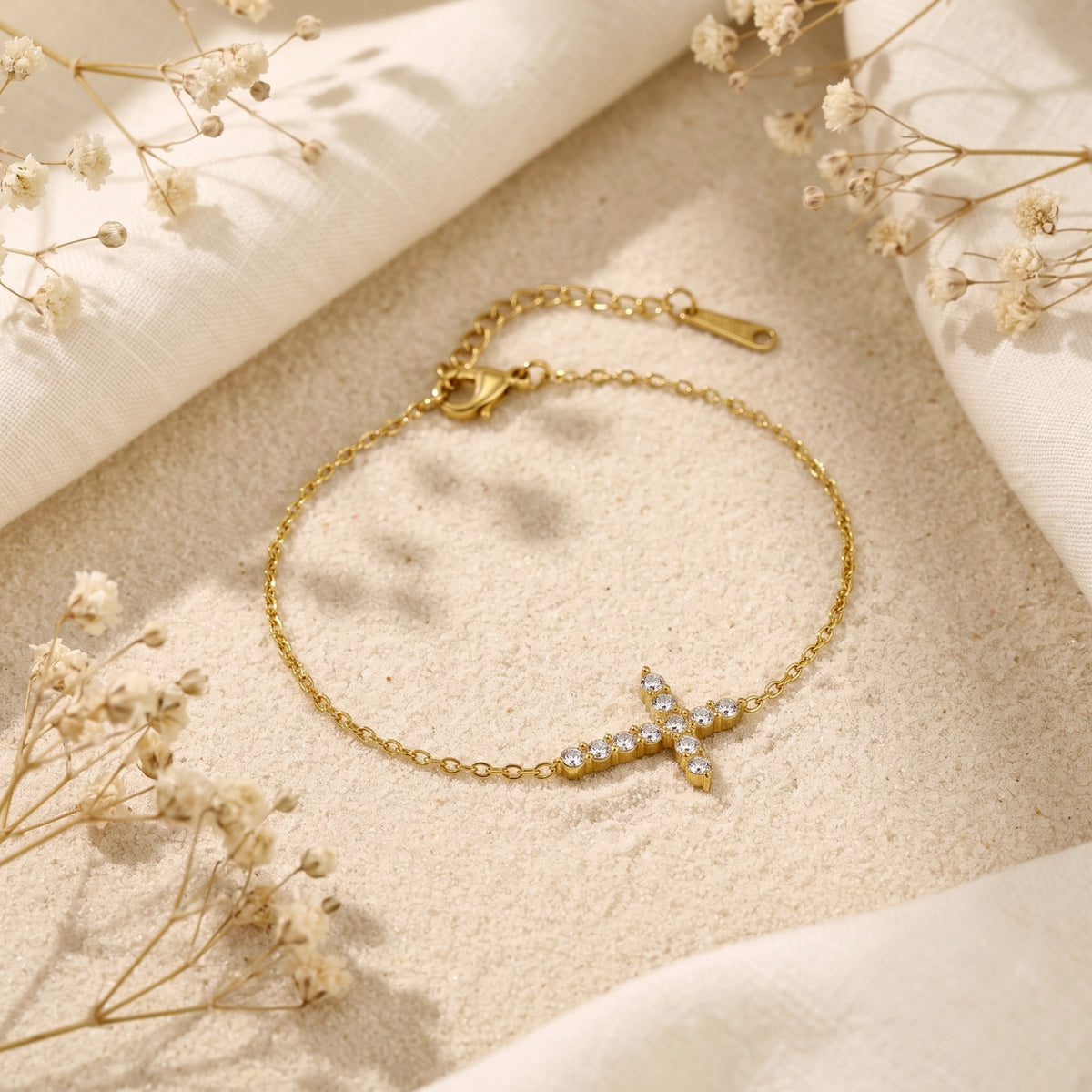 Flat lay view of the gold-tone sideways cross bracelet on sand, showing the adjustable extender chain and clasp.