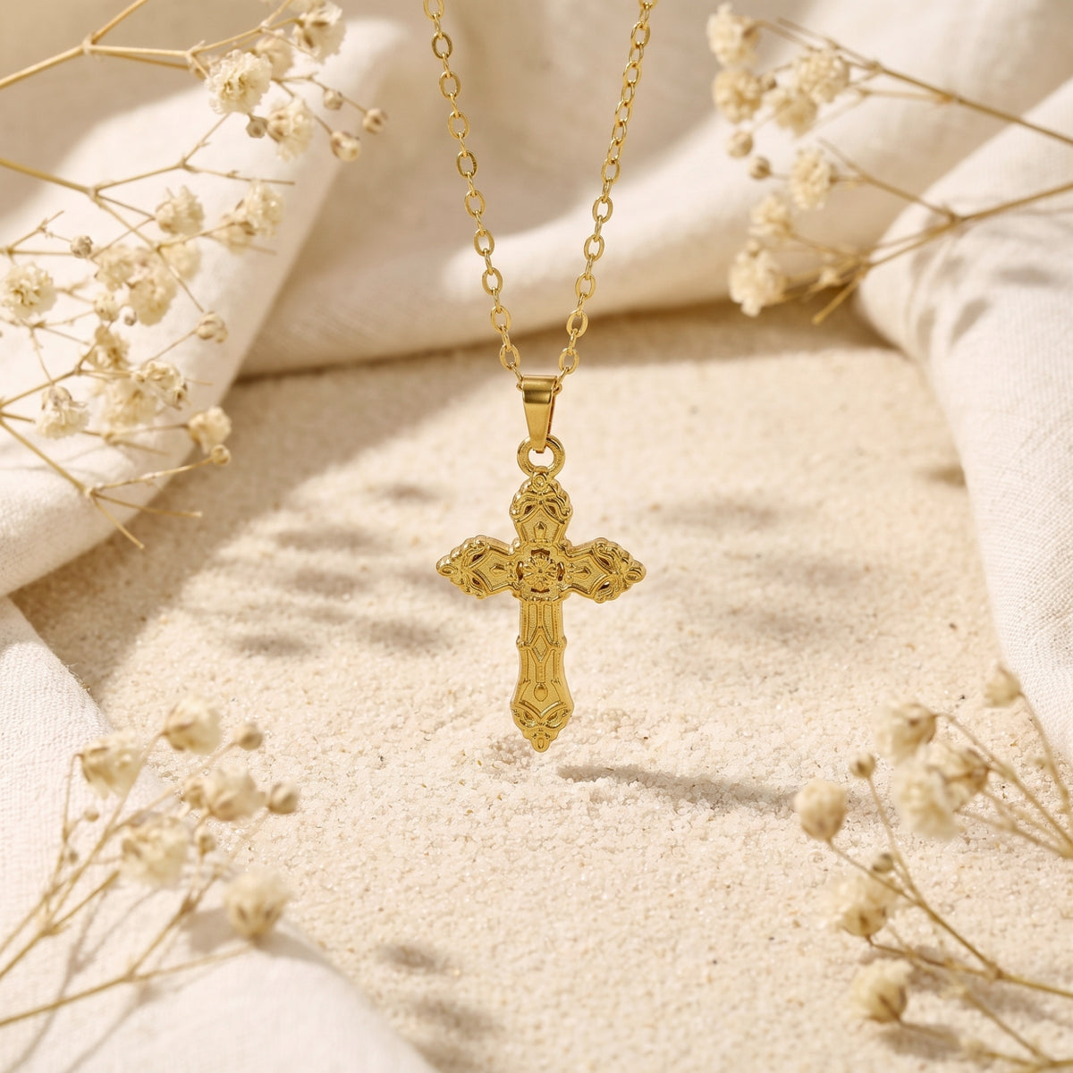 Gold-tone ornate cross pendant hanging above sand, framed by white linen and dried flowers.
