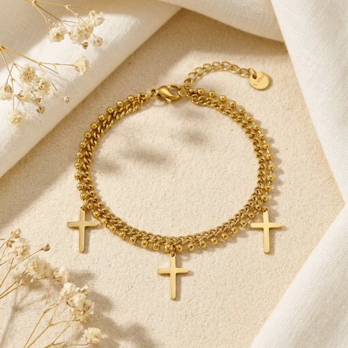 Gold double-strand cross charm bracelet arranged in a circle on a beige surface, showing three cross charms and the clasp.