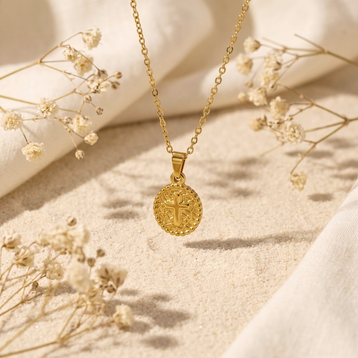 Gold-tone cross medallion necklace suspended above sand, surrounded by dried flowers and white fabric