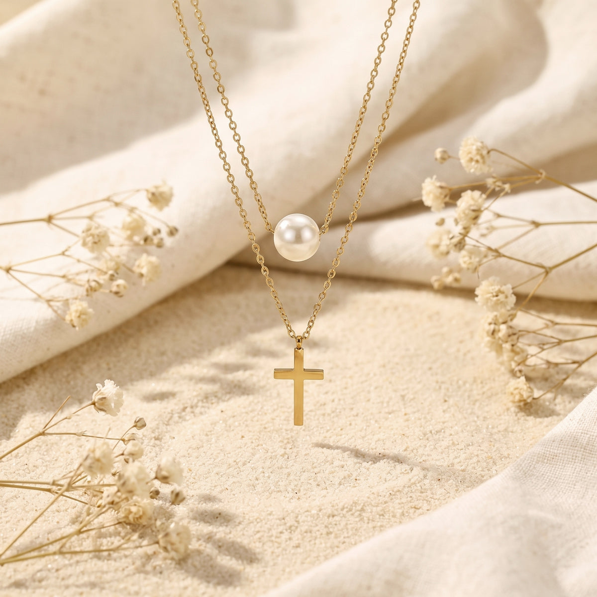 Gold-tone layered necklace shown vertically over sand, with the pearl station above the hanging cross pendant.
