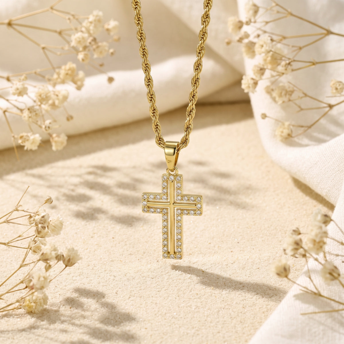 Gold-tone cross necklace suspended above sand with dried flowers, shown in a vertical product view.