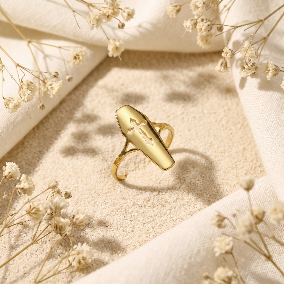 Angled view of a gold-tone coffin ring with a cutout cross tilted on sand among dried flowers.
