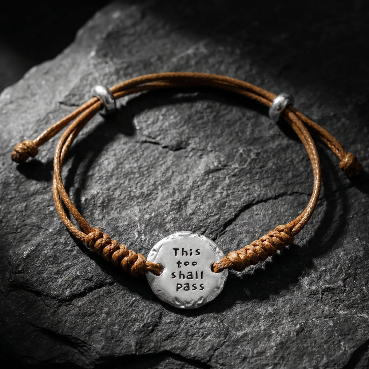 Brown cord bracelet on dark slate with a hammered-edge silver medallion engraved “THIS TOO SHALL PASS.”