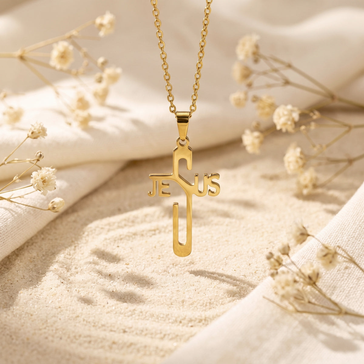 Gold-tone Jesus cross pendant suspended above sand with dried flowers and fabric in the background.