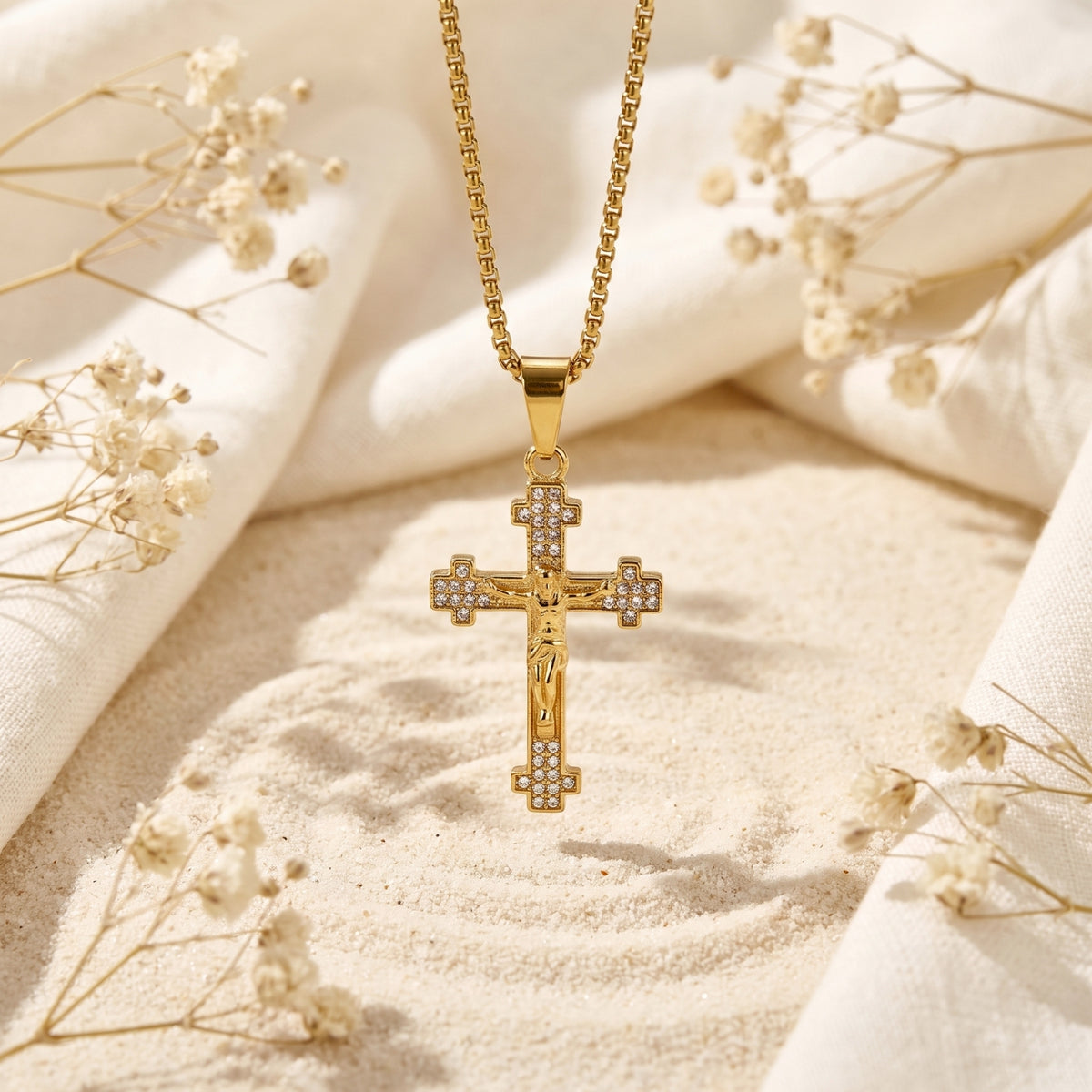 Gold crucifix pendant with crystal accents hanging over sand between draped white fabric and dried flowers.