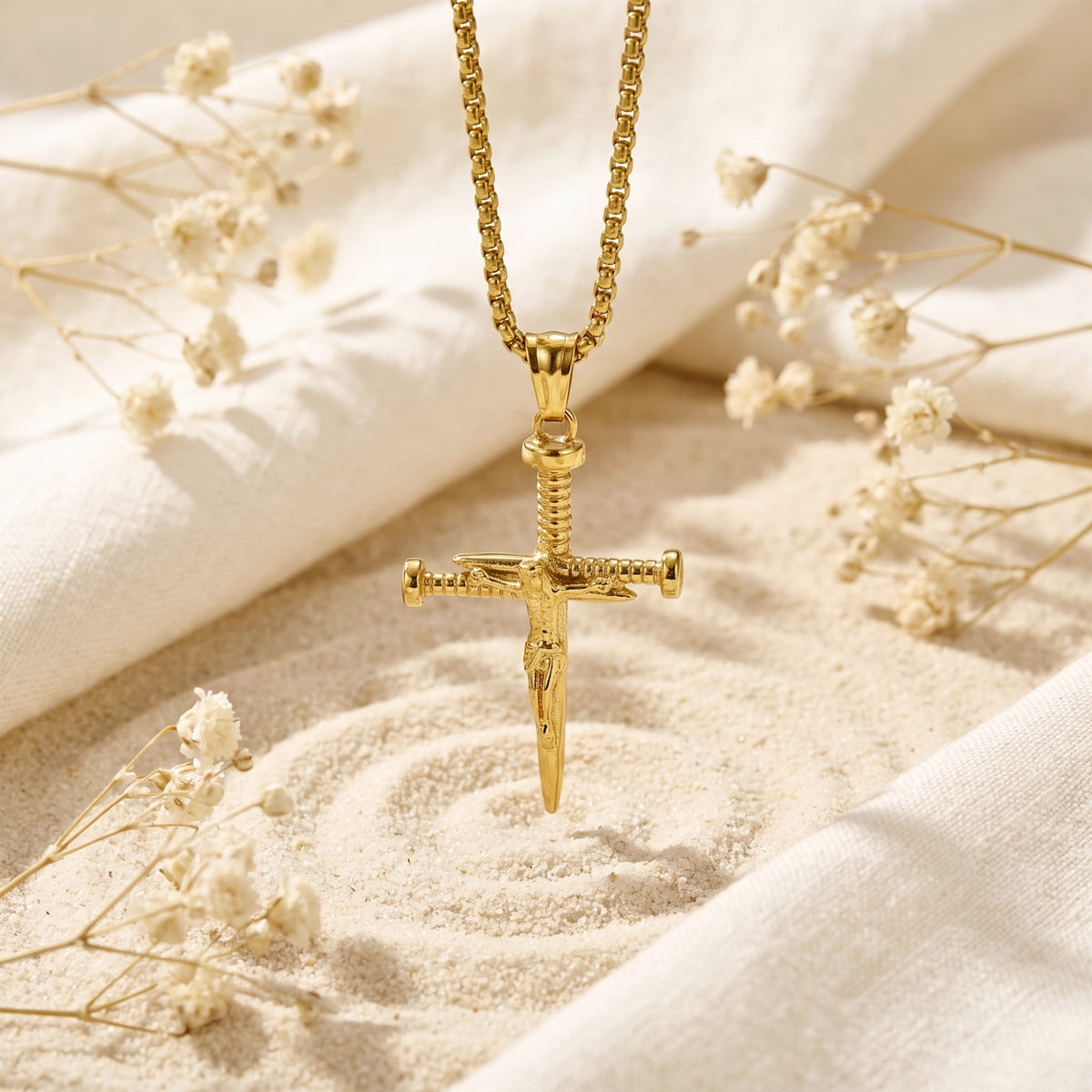 Gold-tone nail cross pendant hanging vertically over raked sand, framed by fabric and dried flowers.