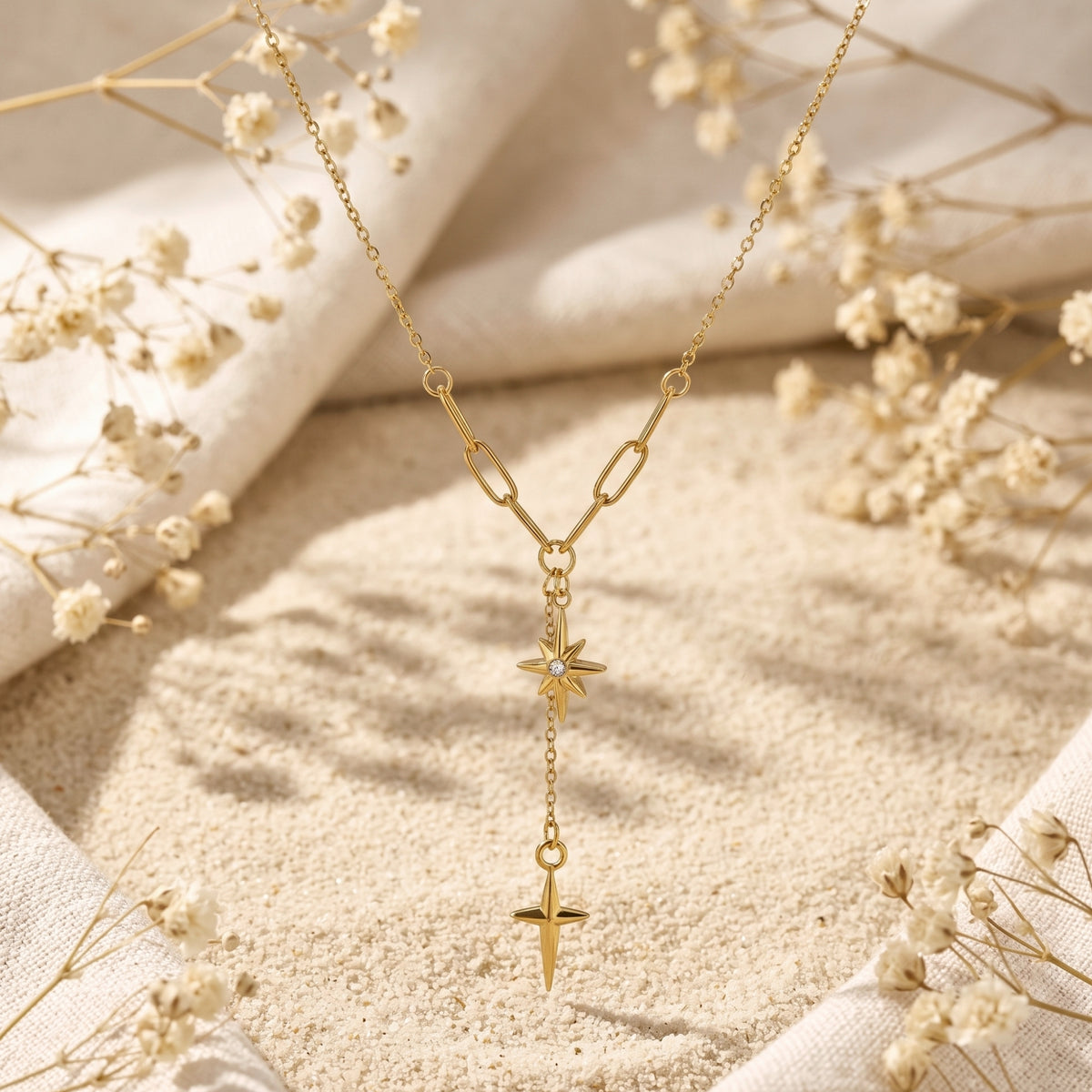 Vertical flat lay of the gold-tone starburst lariat necklace on sand, bordered by white fabric and dried flowers.