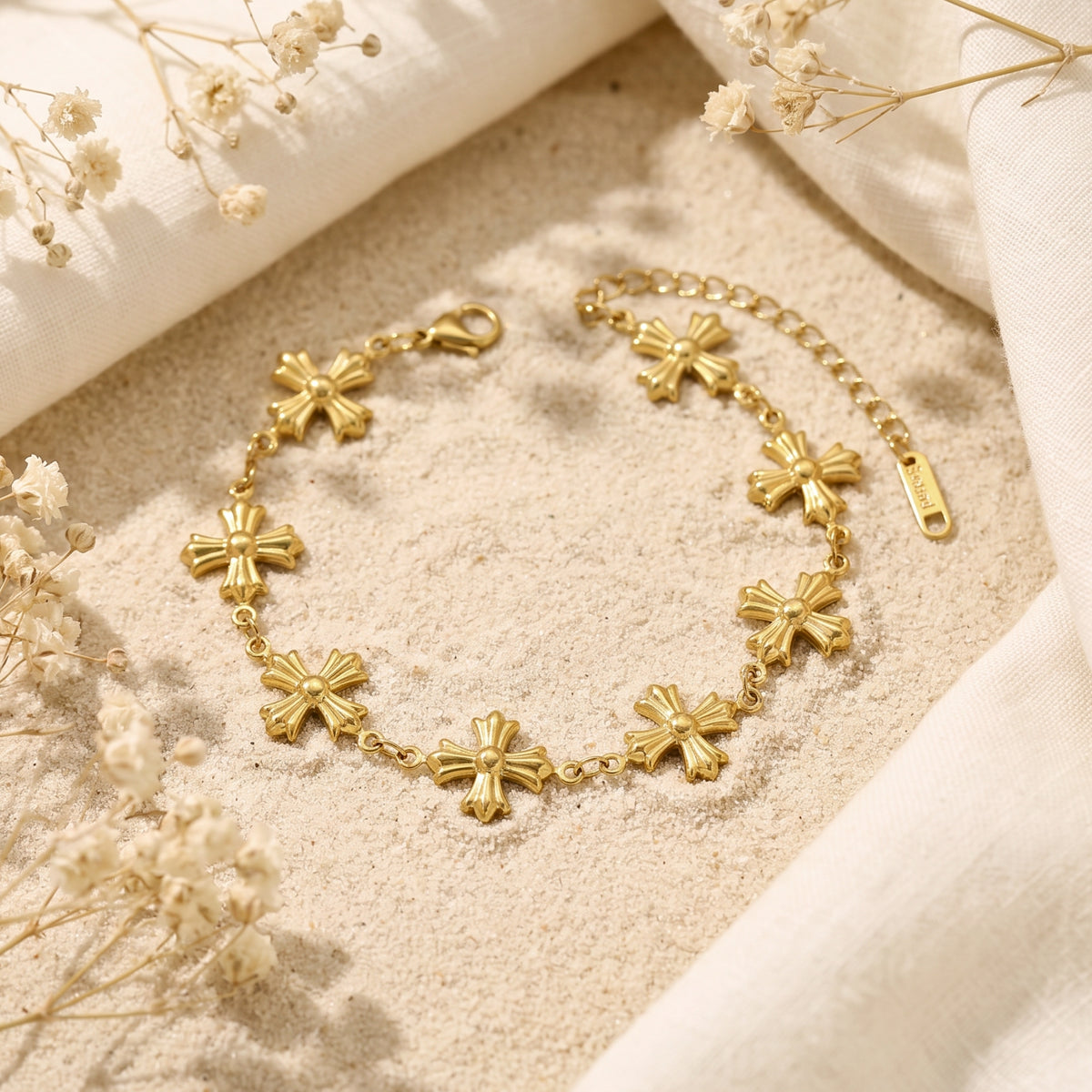 Gold cross station bracelet arranged diagonally on sand beside soft white fabric and dried flowers.
