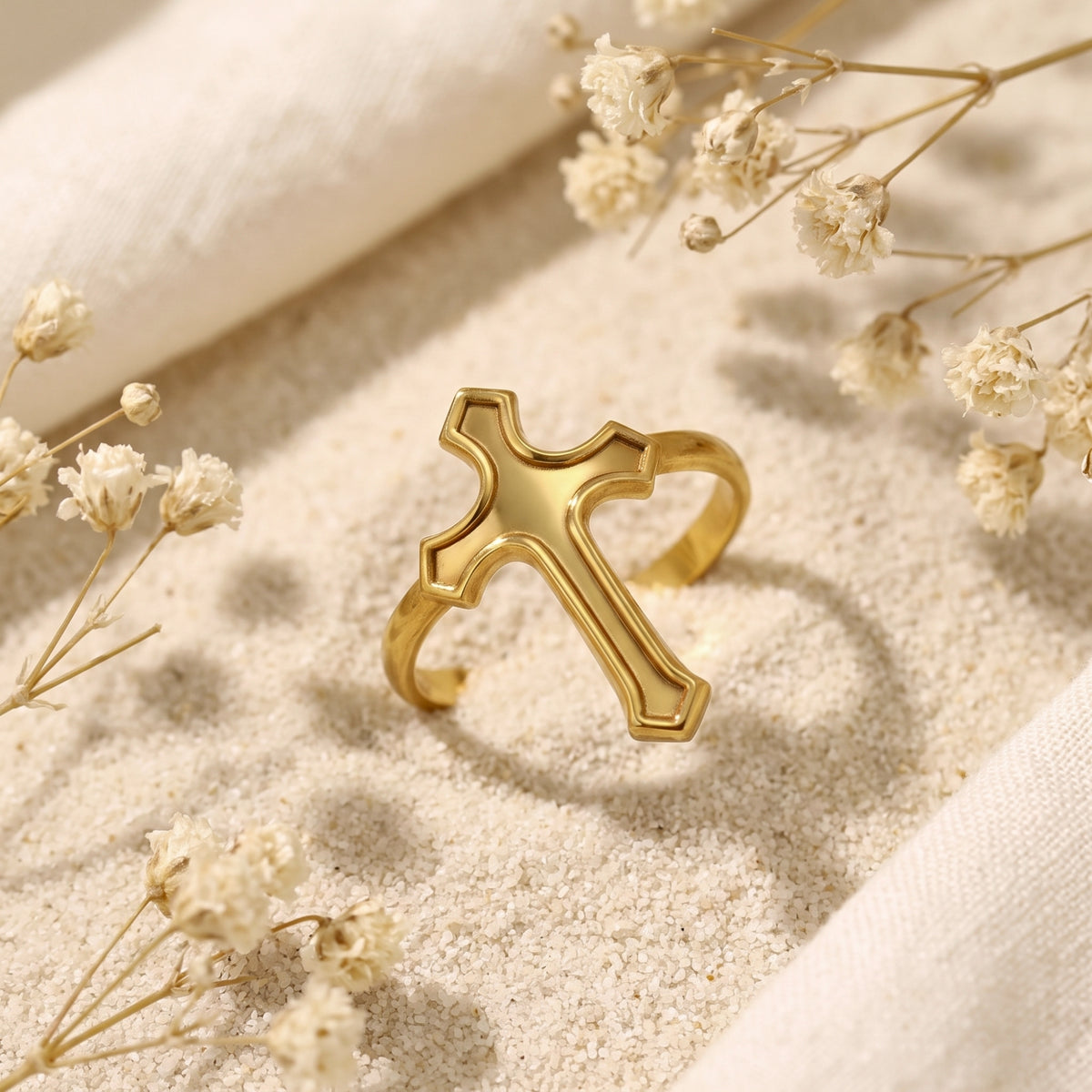Angled view of the gold-tone cross ring displayed on sand beside dried flowers.