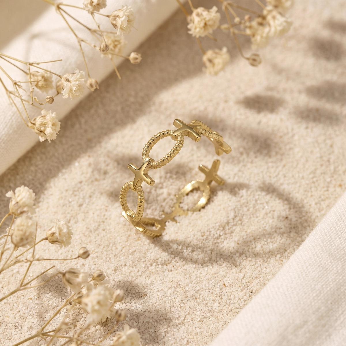 Angled view of a gold-tone open cross ring propped on sand among dried white flowers.