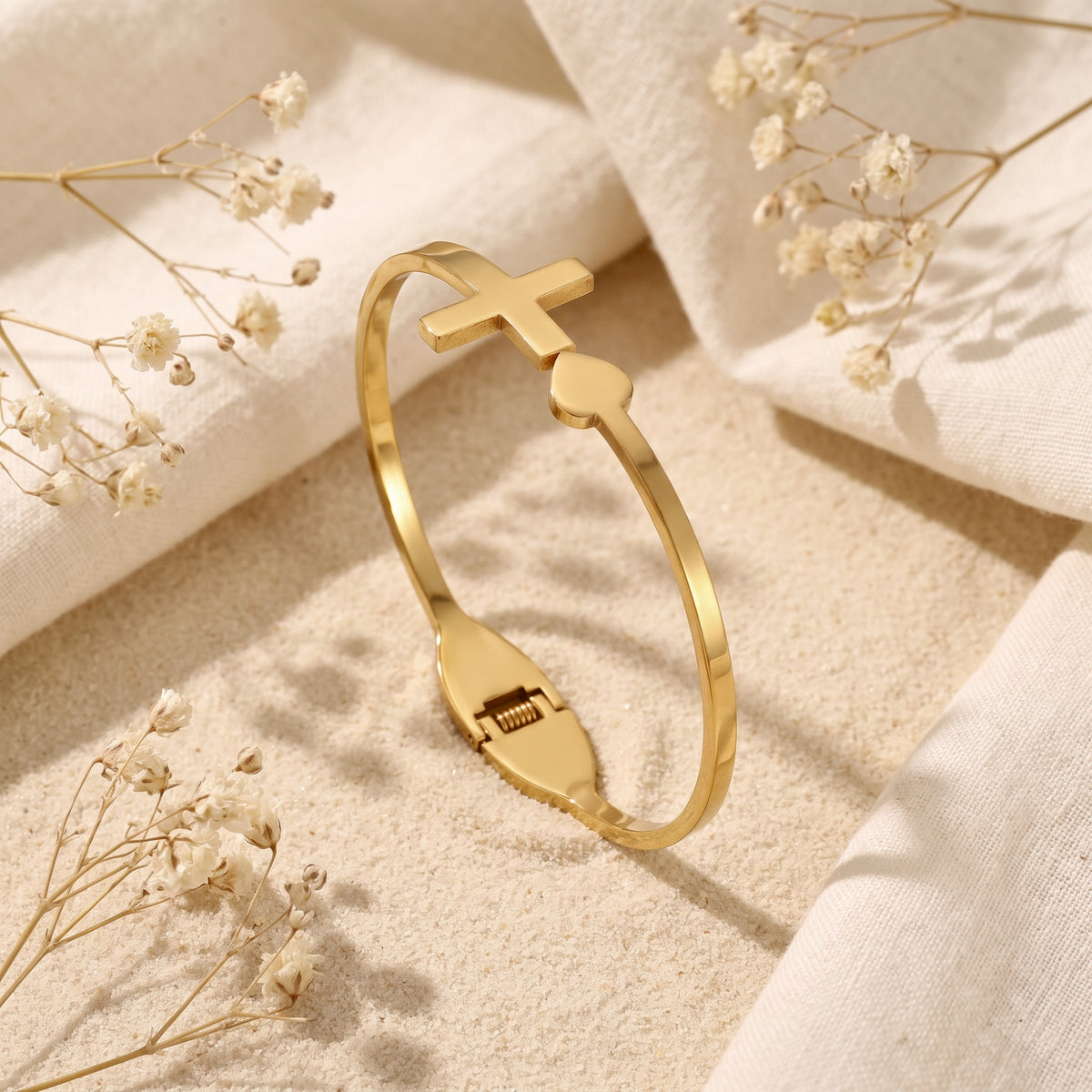 Gold-tone cross and heart hinged bangle displayed upright and open on textured sand with dried flowers.