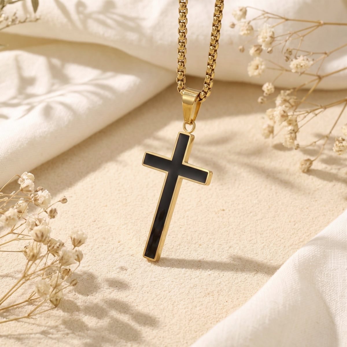 Gold-tone cross pendant with black enamel inlay hanging from a box chain above sand and dried flowers.