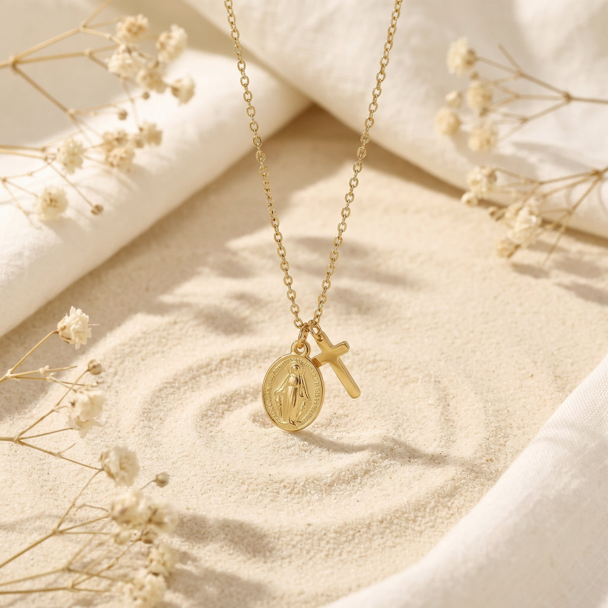 Gold-tone cross and oval medal necklace hanging over a sand display with dried flowers.