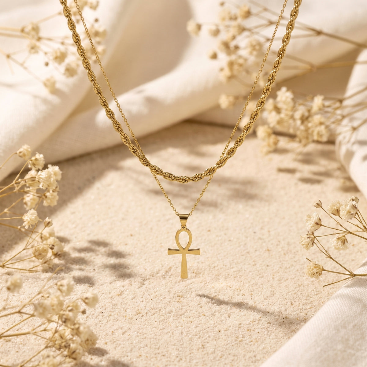 Layered gold-tone Ankh pendant necklace hanging above a sandy surface with dried white flowers.