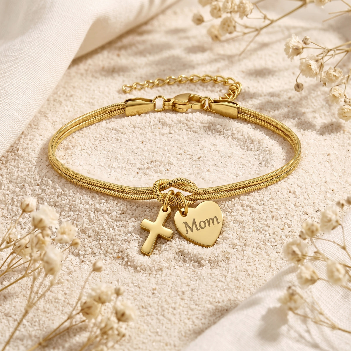 Flat display of the gold-tone knot bracelet on sand, showing its clasp, cross, and engraved “Mom” heart charm.