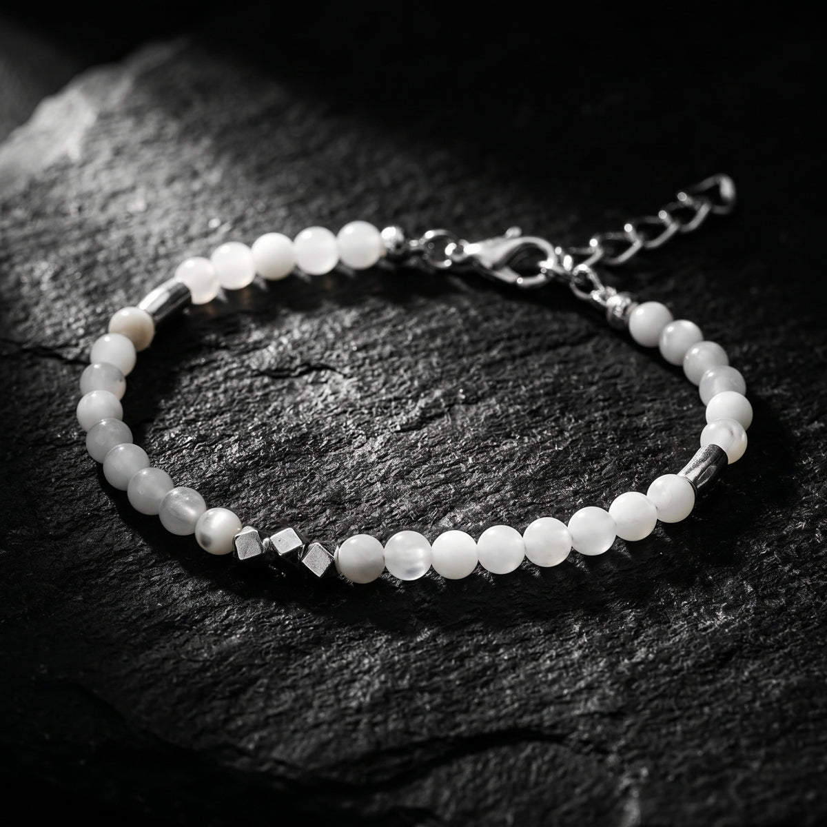 Angled close-up of the white beaded bracelet with silver cube accents and extension chain on textured dark slate.