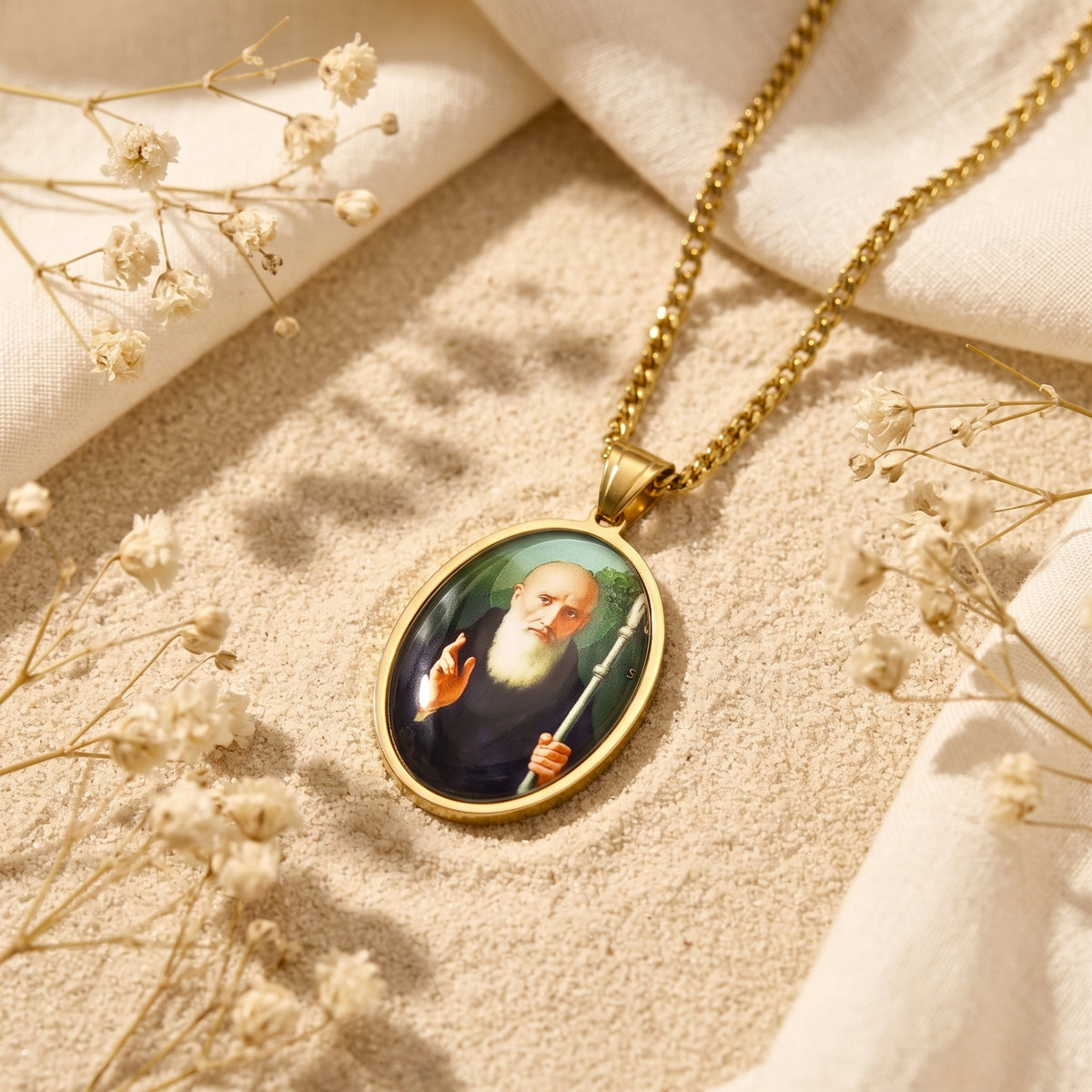 Gold-tone oval saint medallion pendant resting at an angle on sand beside white cloth and dried flowers.