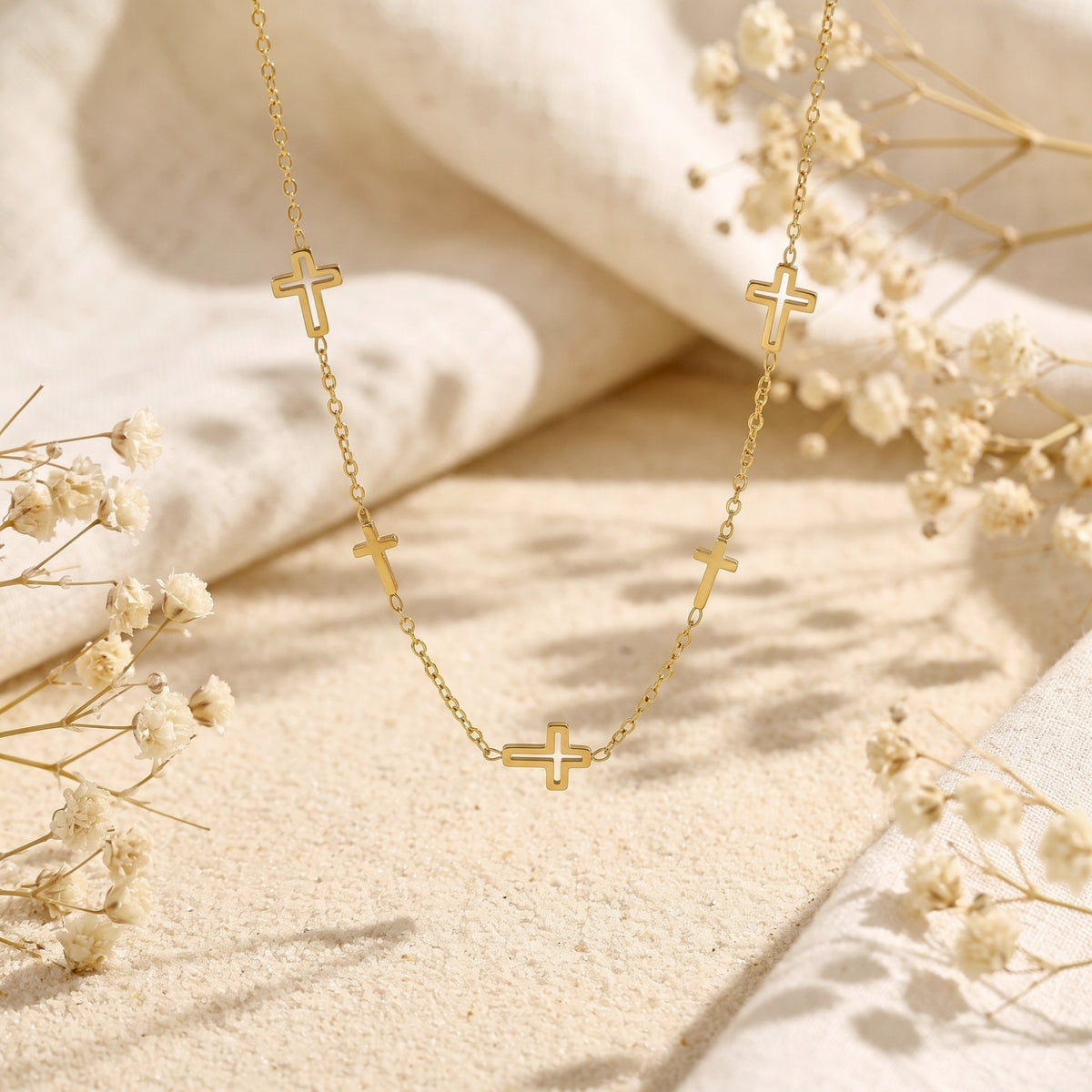 Gold-tone multi-cross necklace arranged in a U shape on a sandy surface with white fabric and dried flowers.