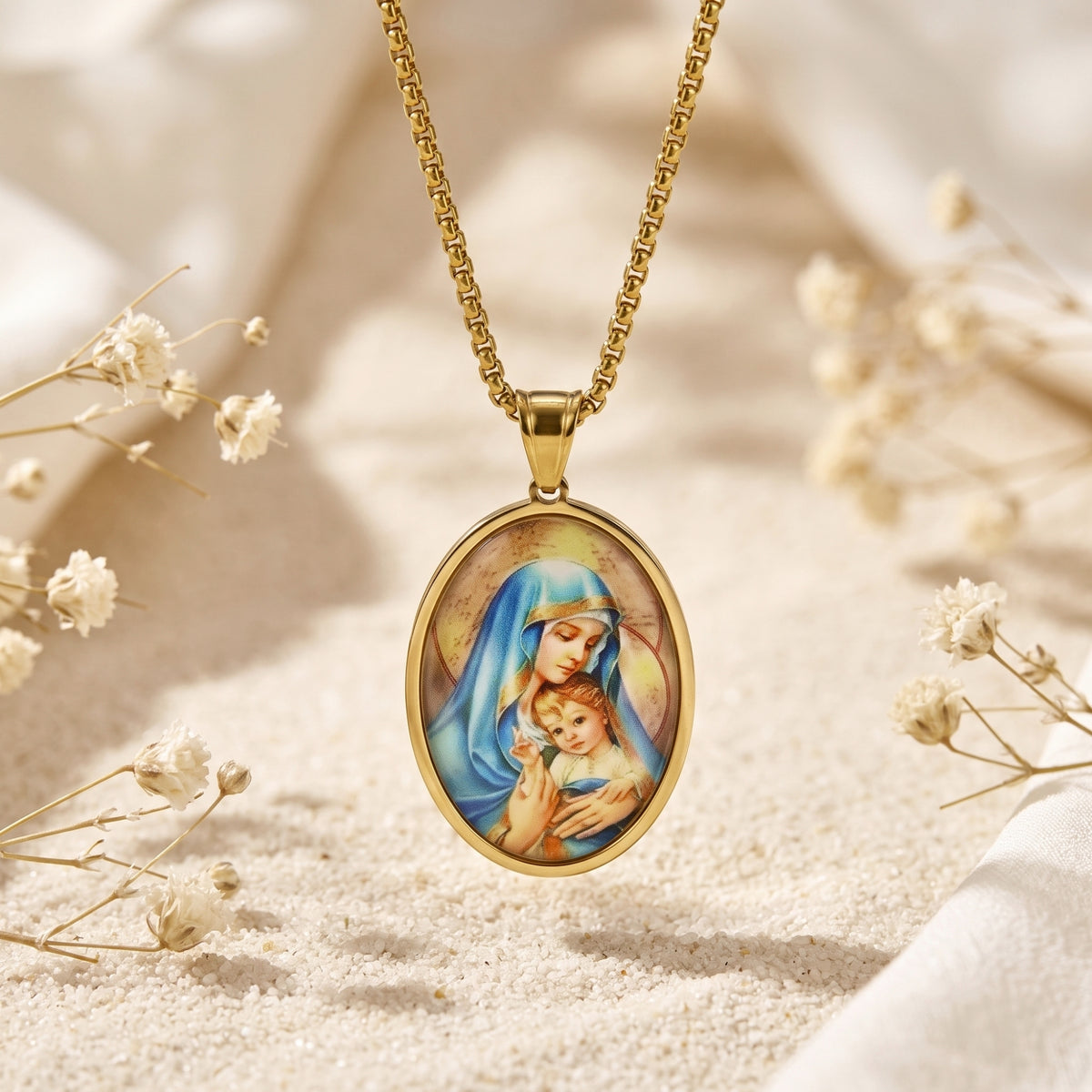 Front view of the gold-tone oval Madonna and Child pendant hanging over sand and white flowers.