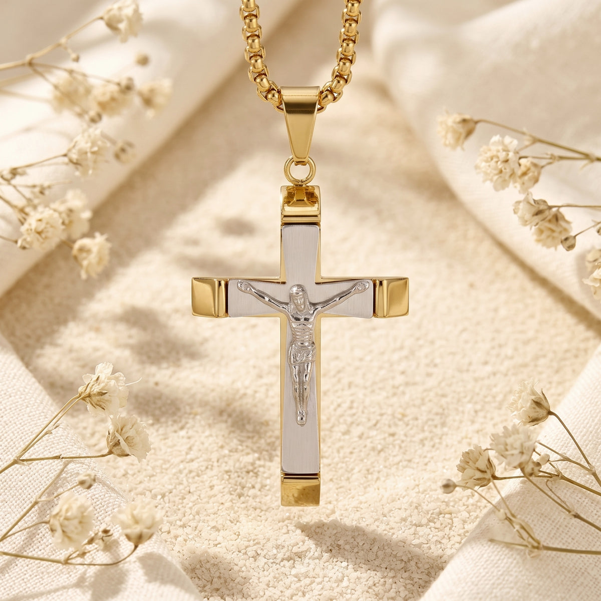 Layered crucifix pendant hanging vertically from its gold-tone chain against sand and dried flowers.