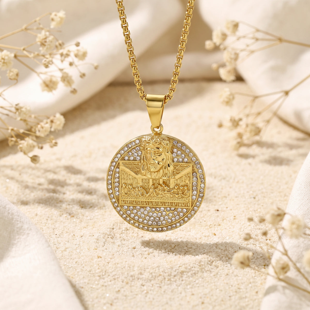 Frontal gold-tone Last Supper medallion pendant with crystal accents hanging above sand and white cloth.