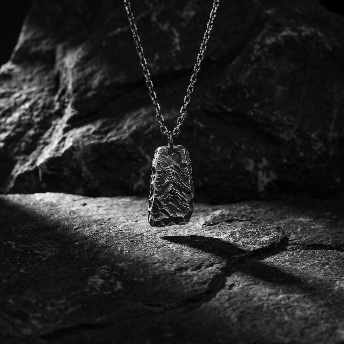 Textured silver ridge pendant hanging vertically against a dark slate rock background.