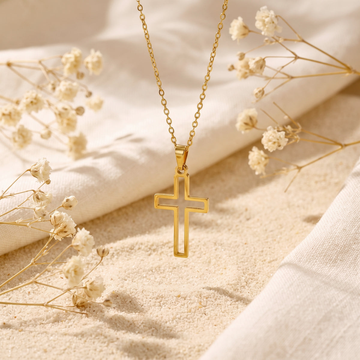 Gold-tone cutout cross pendant hanging vertically above textured sand, white fabric, and dried flowers.