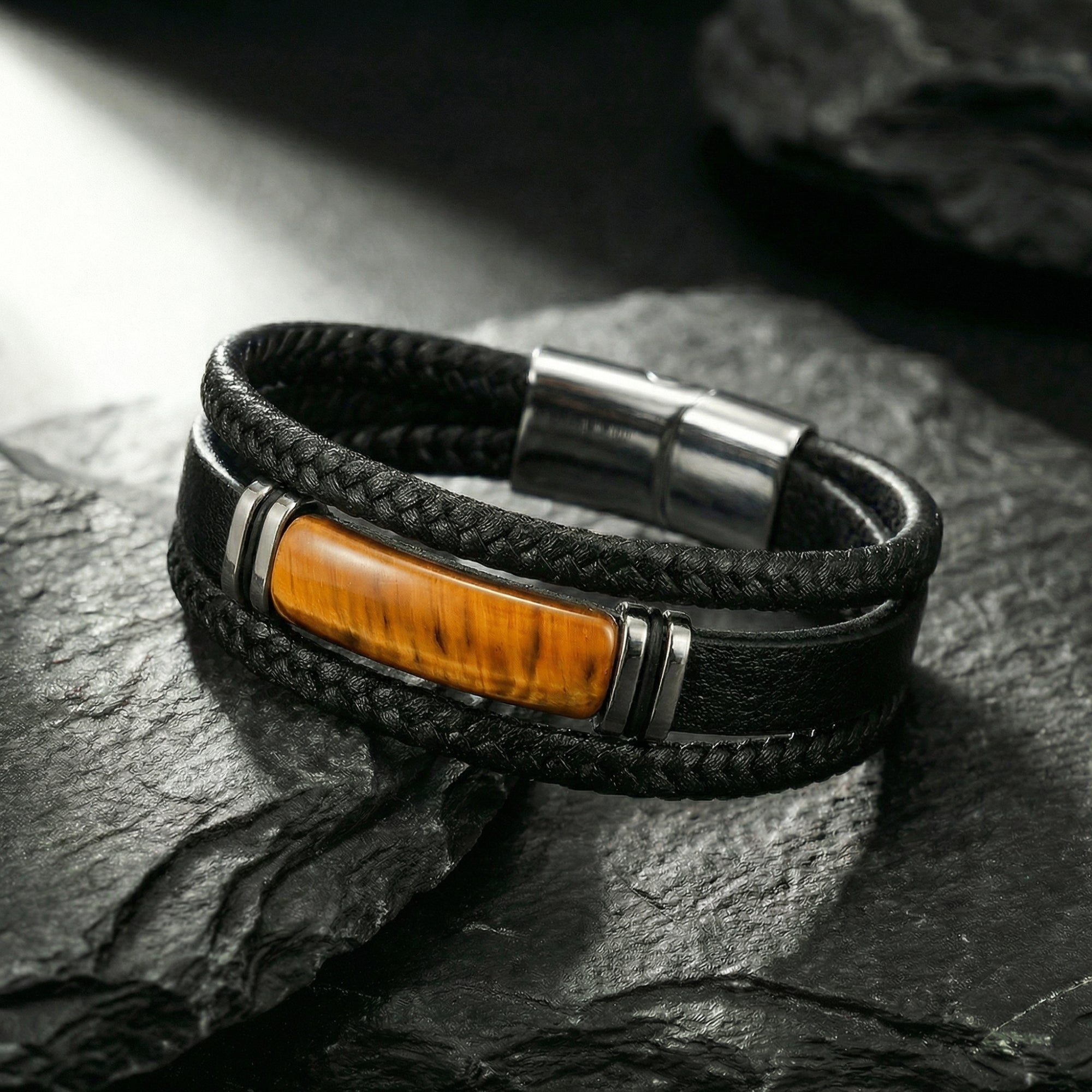 The Forge braided leather bracelet with tiger's eye stone and black steel magnetic clasp on a dark rock surface.