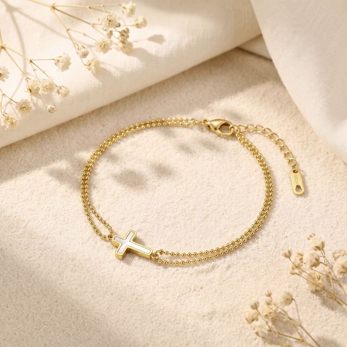 Top-down view of the gold double-bead cross bracelet on fabric, showing its extender chain and dried flowers.