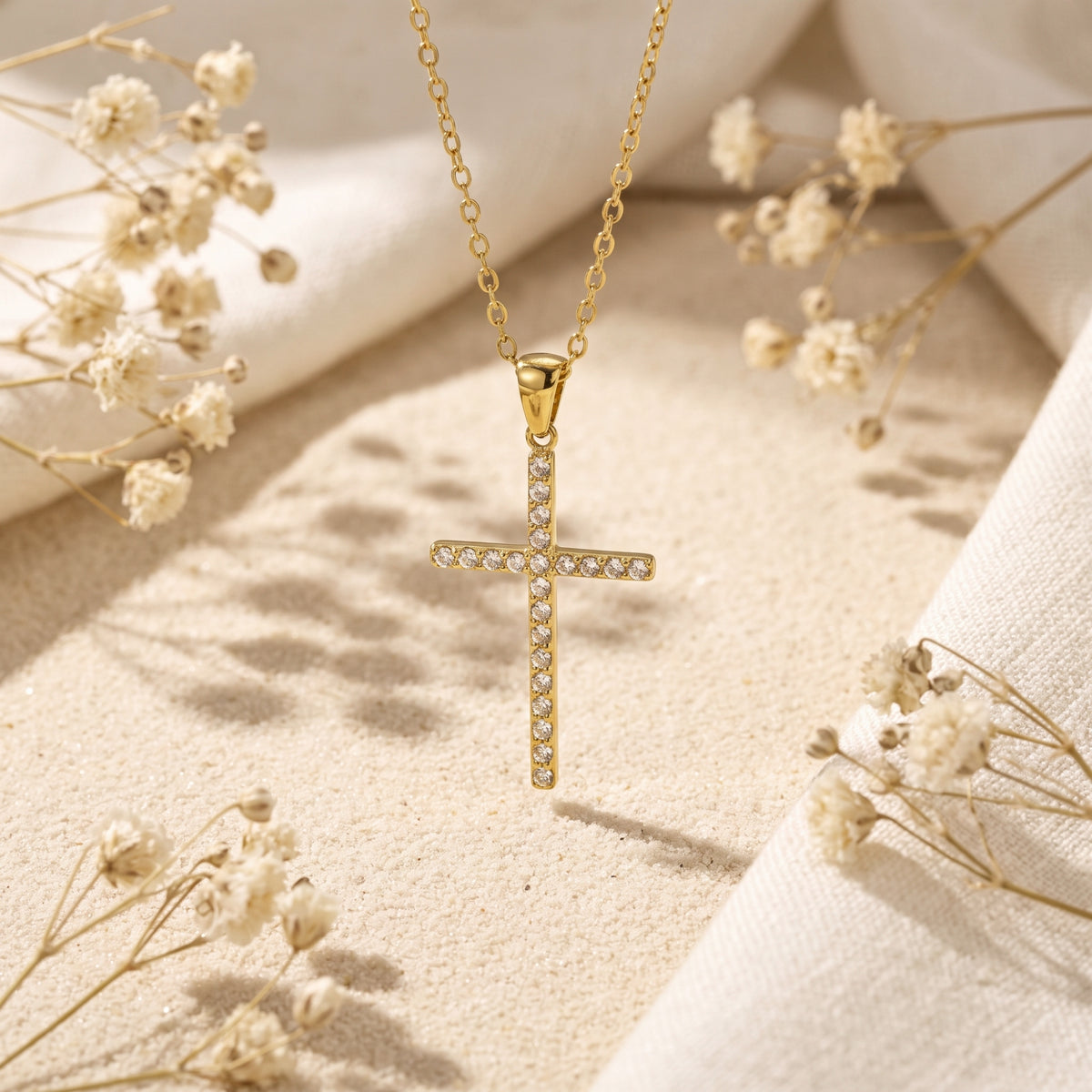 Gold-tone pave cross pendant hanging above pale sand, surrounded by dried white flowers.