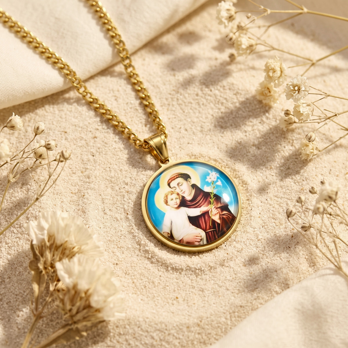 Close-up of a gold-tone Saint Anthony medallion necklace lying on sand among white dried flowers.