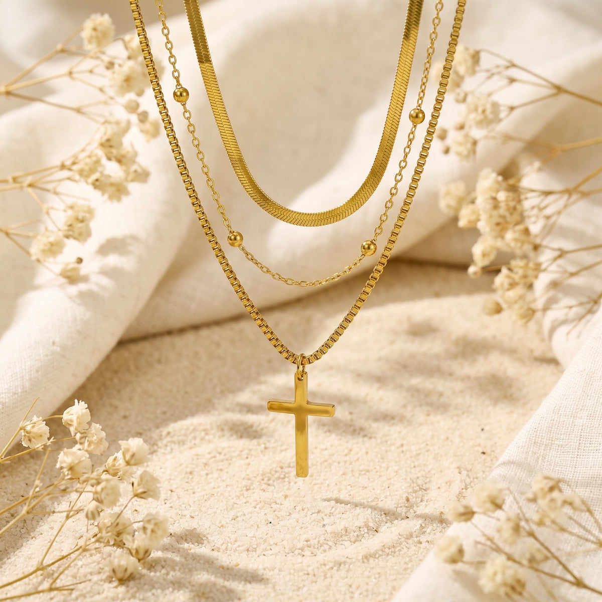 Gold-tone triple-layered cross necklace hanging over sand, white fabric, and dried flowers.