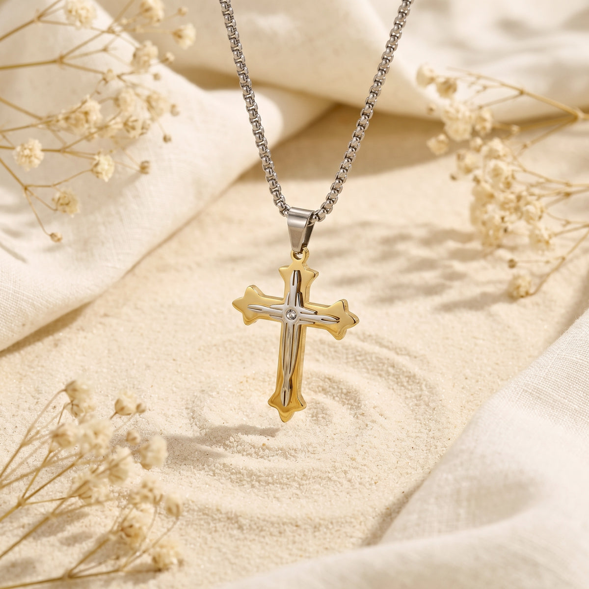 Close view of the two-tone cross pendant lying on a sand-colored surface, with its chain extending upward.