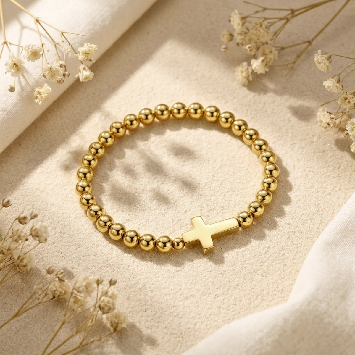 Angled view of a gold-tone beaded cross bracelet resting on textured sand with dried white flowers.