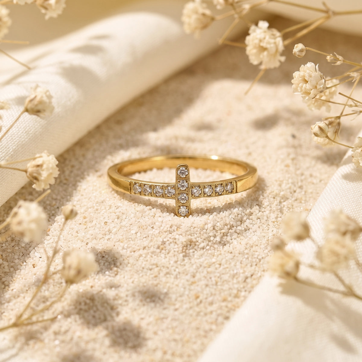 Front-facing gold-tone cross ring with rows of clear crystals displayed horizontally on pale sand.