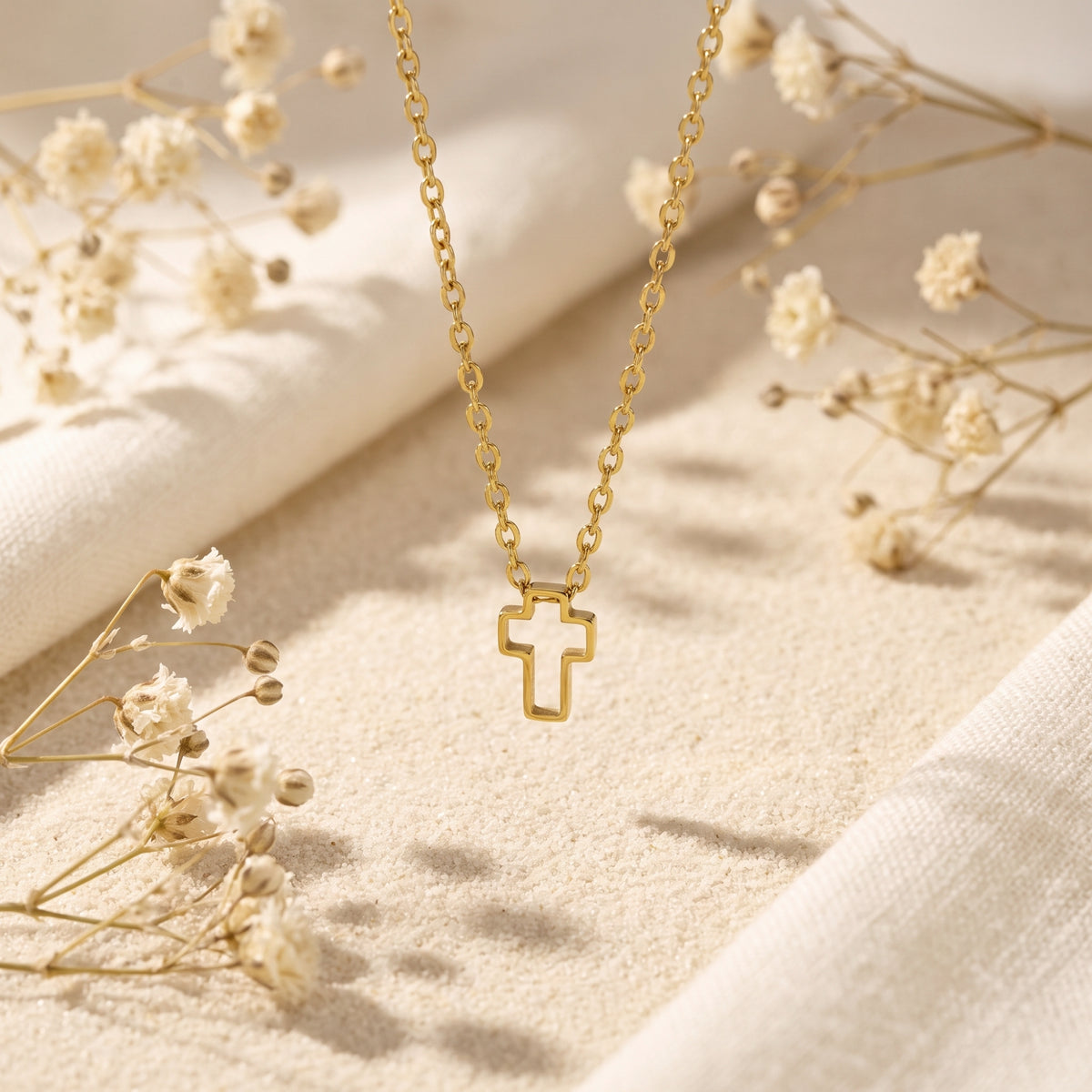 Gold-tone openwork cross necklace hanging above sand, with white fabric and dried flowers in the background.