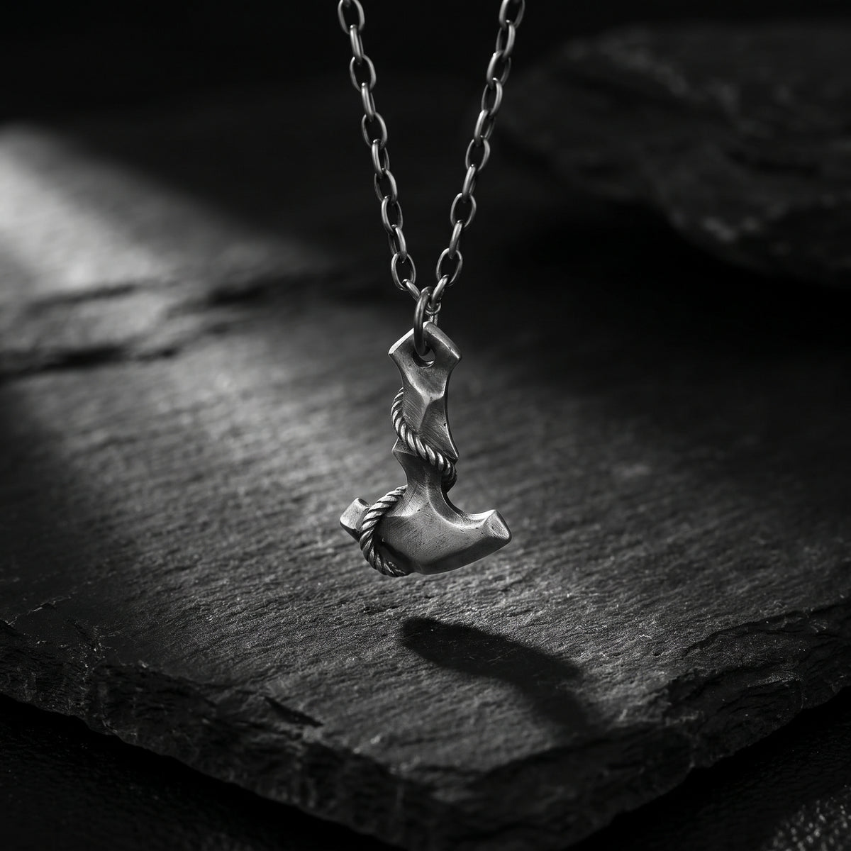 Silver Mjolnir hammer pendant hanging vertically above dark slate in direct light.