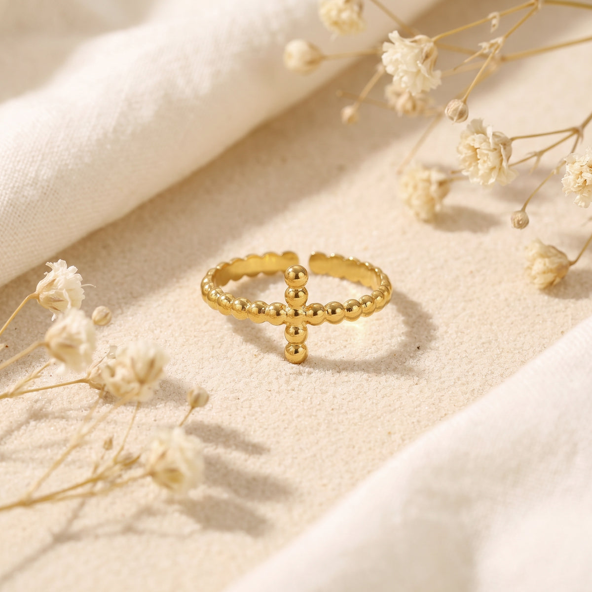 Front view of a gold-toned beaded cross ring on sand, showing its beaded cross motif and open band.
