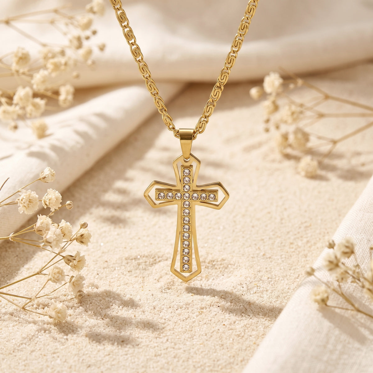 Frontal view of the gold-finish crystal-set cross pendant suspended above sand between dried flowers.