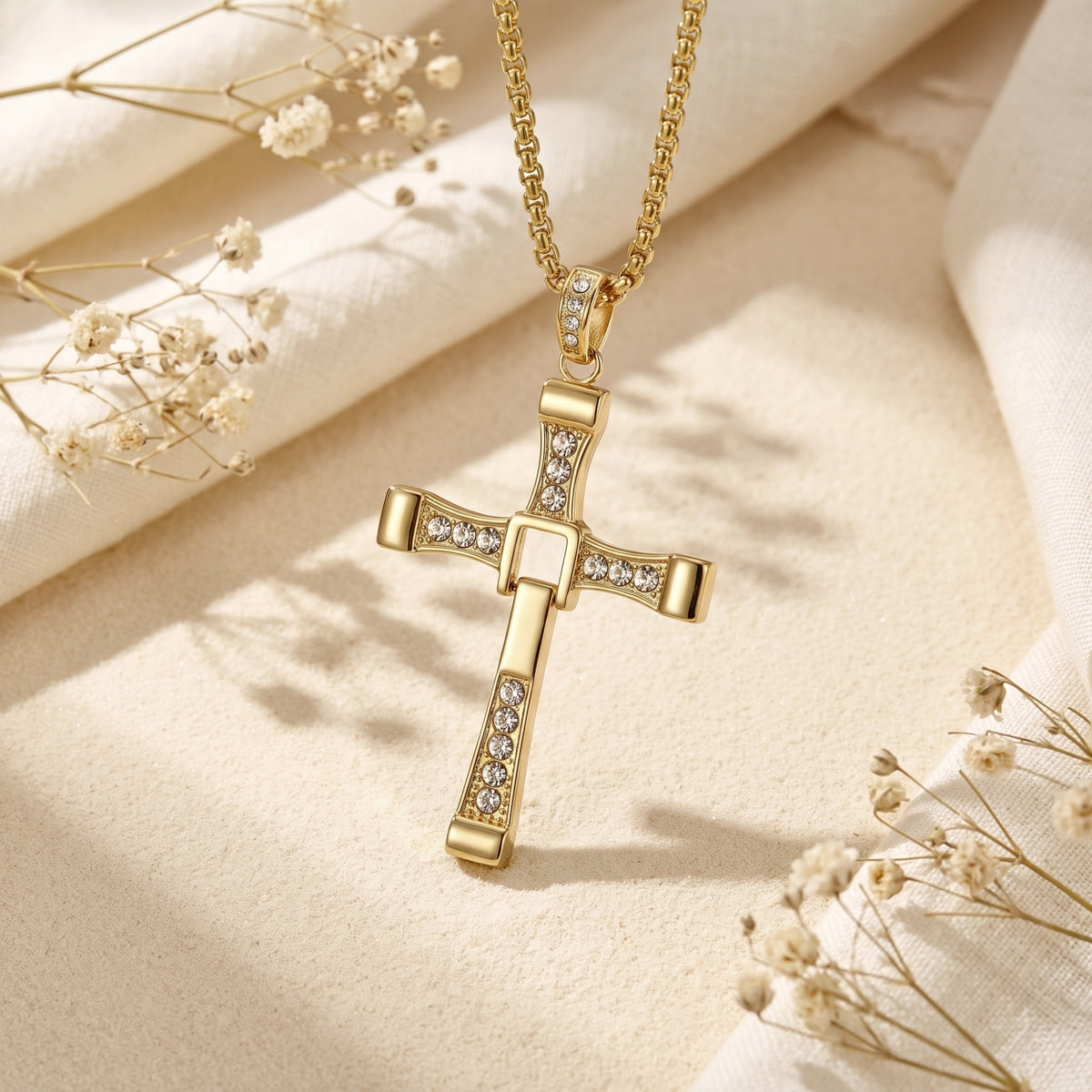 Gold-tone hinged cross pendant with clear crystals hanging upright over sand beside white fabric and dried flowers.