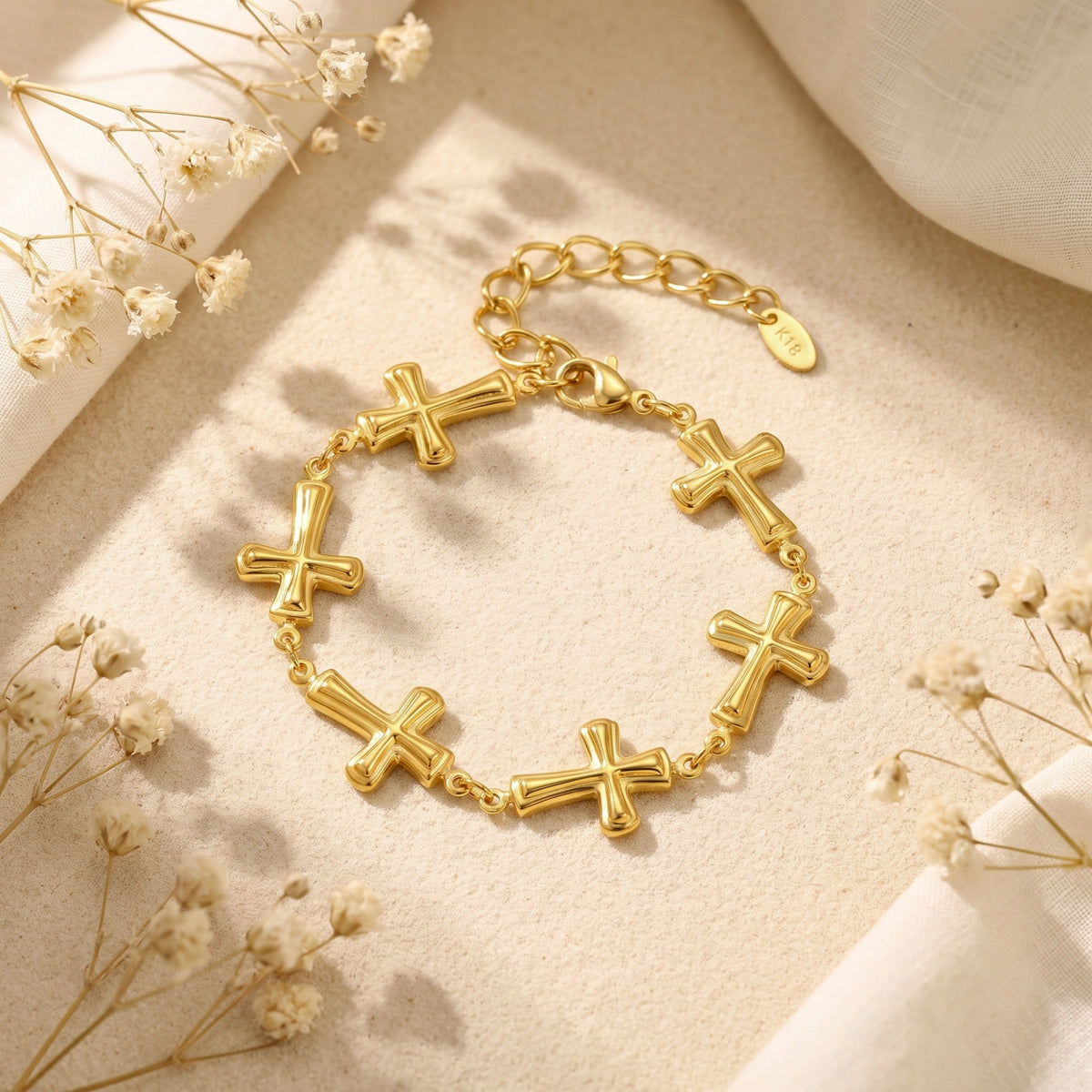 Flat lay view of the gold-tone cross station bracelet arranged in a circle on sand with white dried flowers.