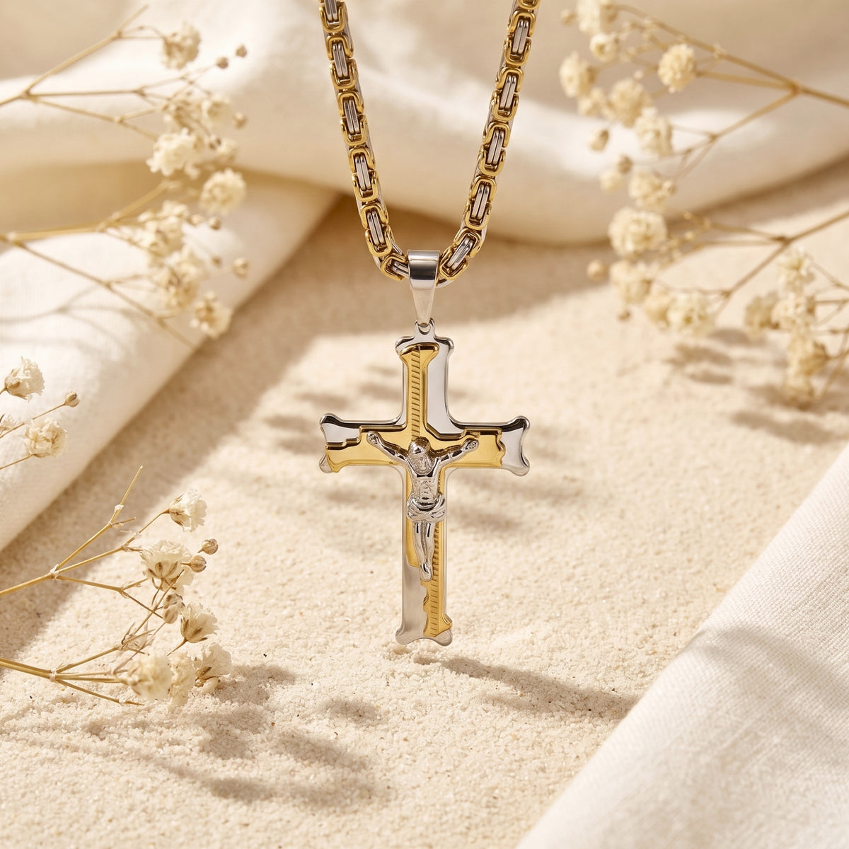 Two-tone crucifix pendant suspended vertically over sand with surrounding dried flowers.