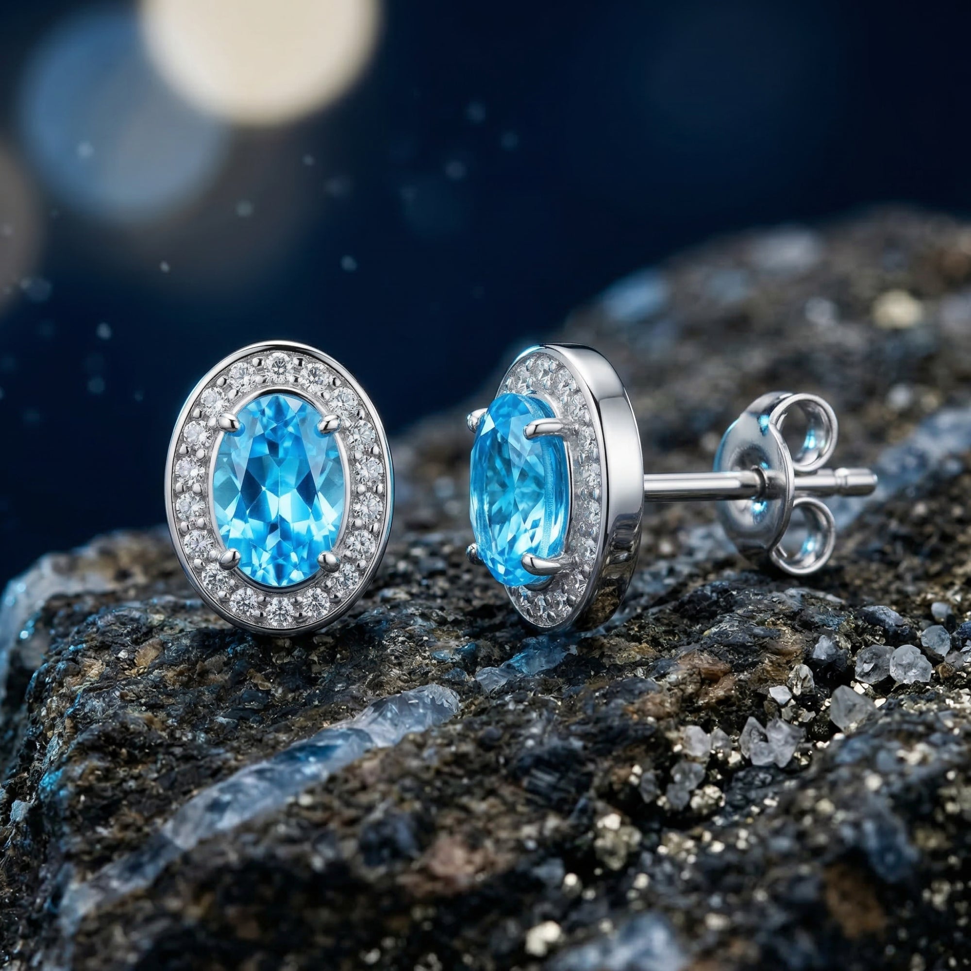 White gold oval natural blue topaz halo stud earrings shown from the front and side on a dark rock.