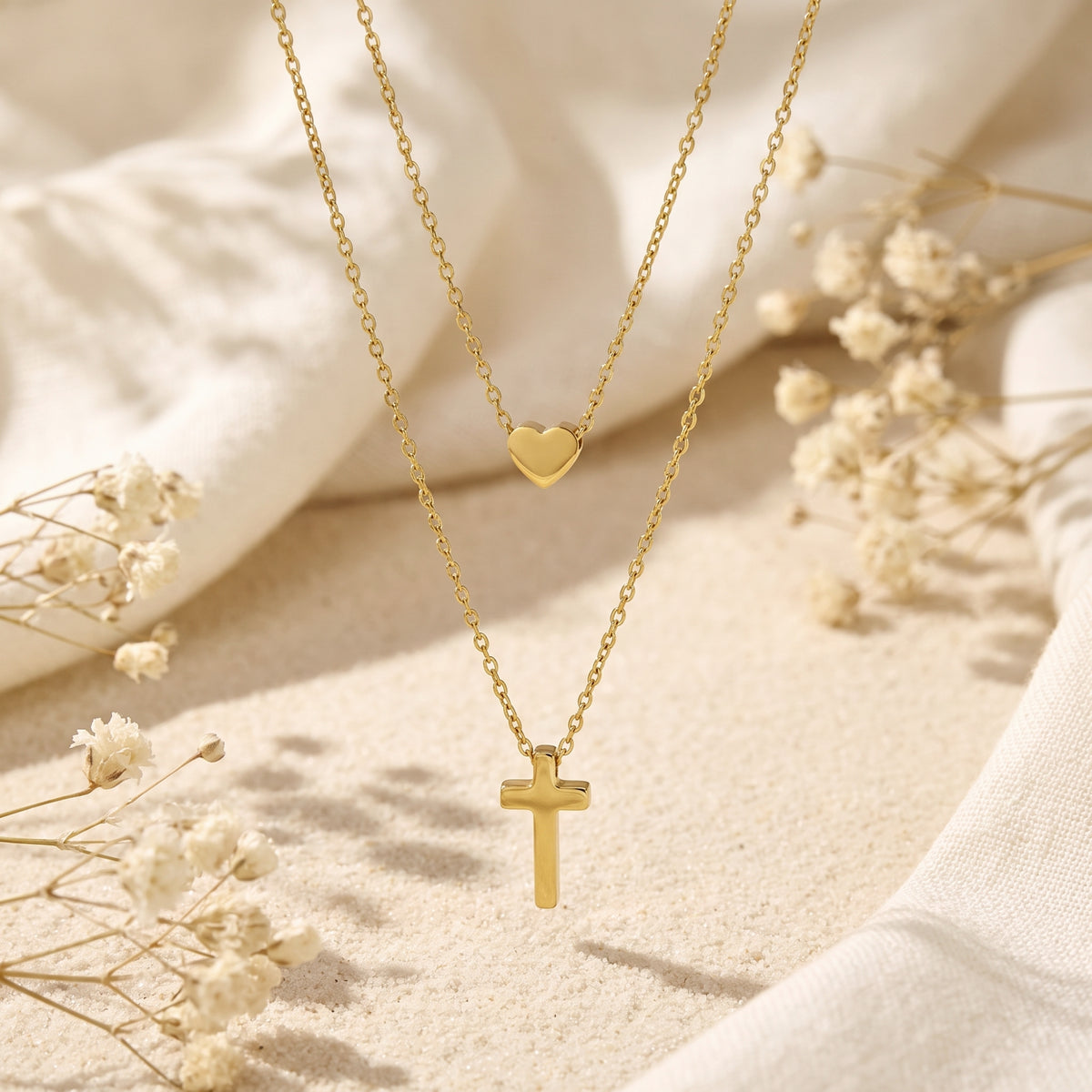 Flat lay of the layered gold-tone necklace with heart and cross pendants arranged vertically on sand.