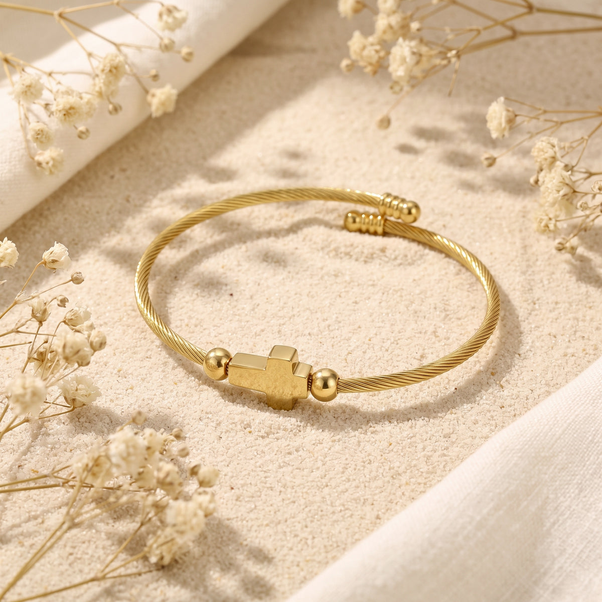 Angled view of the gold-tone cross cable bracelet on sand, showing its open bypass cuff design.