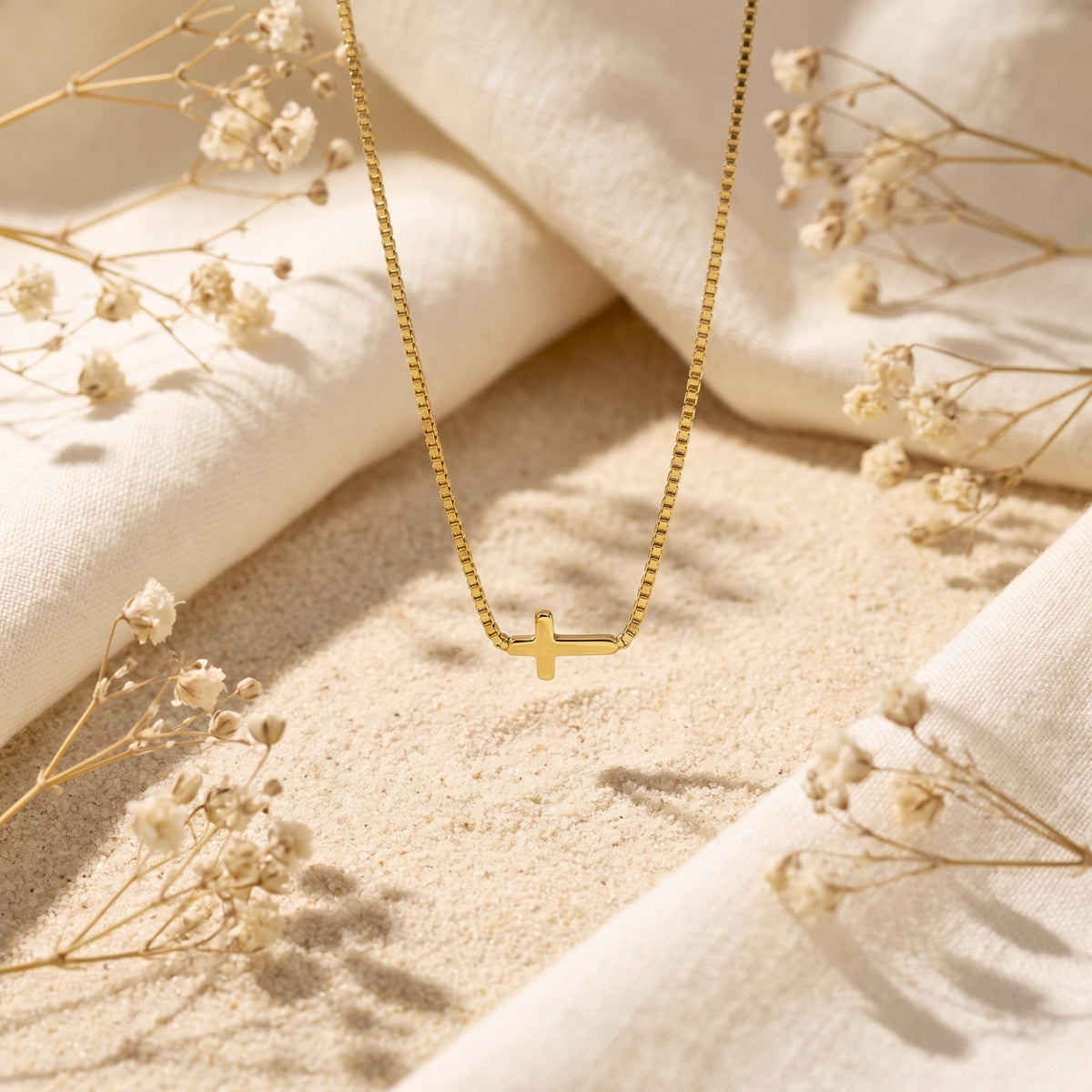 Gold-tone cross necklace laid vertically on pale sand between white fabric and dried flowers.