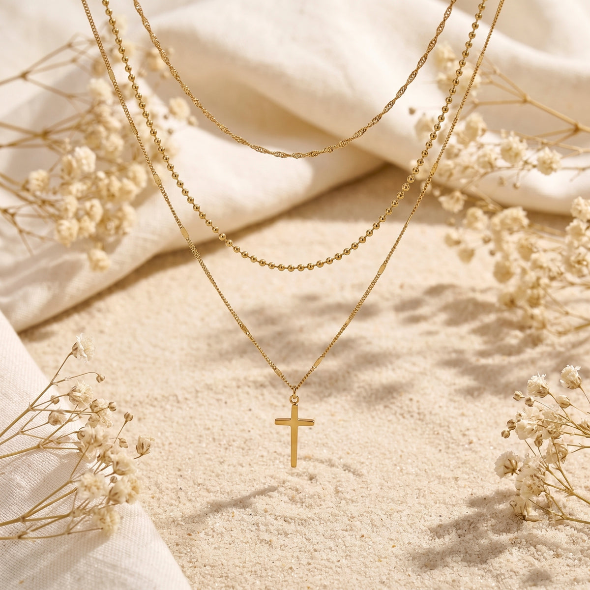 Gold-tone three-strand layered necklace with cross pendant arranged on sand, white fabric, and dried flowers.