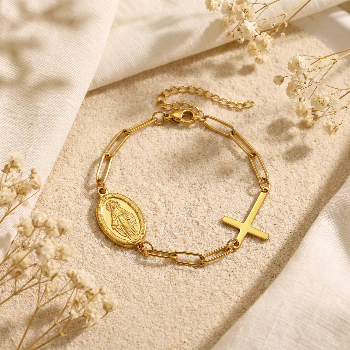 Top-down view of the full gold-tone link bracelet with an oval Virgin Mary medal and cross charm on sand.